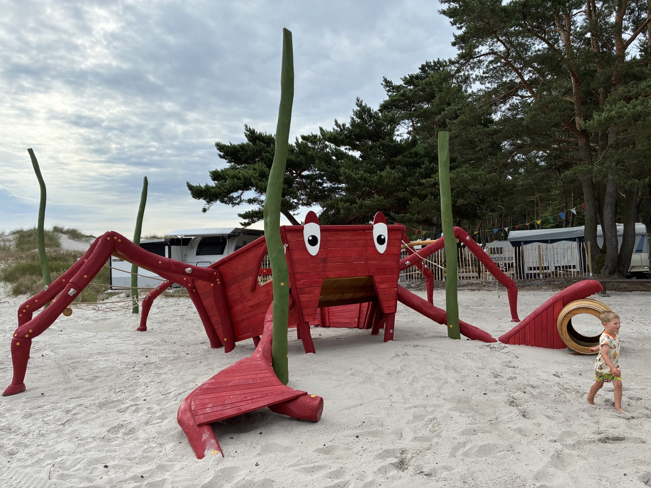 Rote, krabbenförmige Spielplatzanlage auf sandigem Boden, auf der Kinder Spaß und Spiel genießen, daneben ein Kind, das vorbeiläuft, und im Hintergrund Bäume.