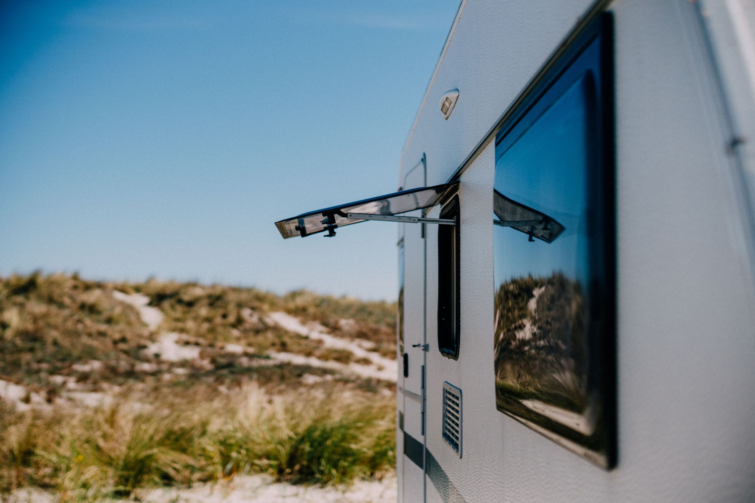 Nahaufnahme eines offenen Wohnmobilfensters mit Blick auf grasbewachsene Dünen und blauen Himmel - genießen Sie die Freiheit der Mietunterkünfte in der Natur.