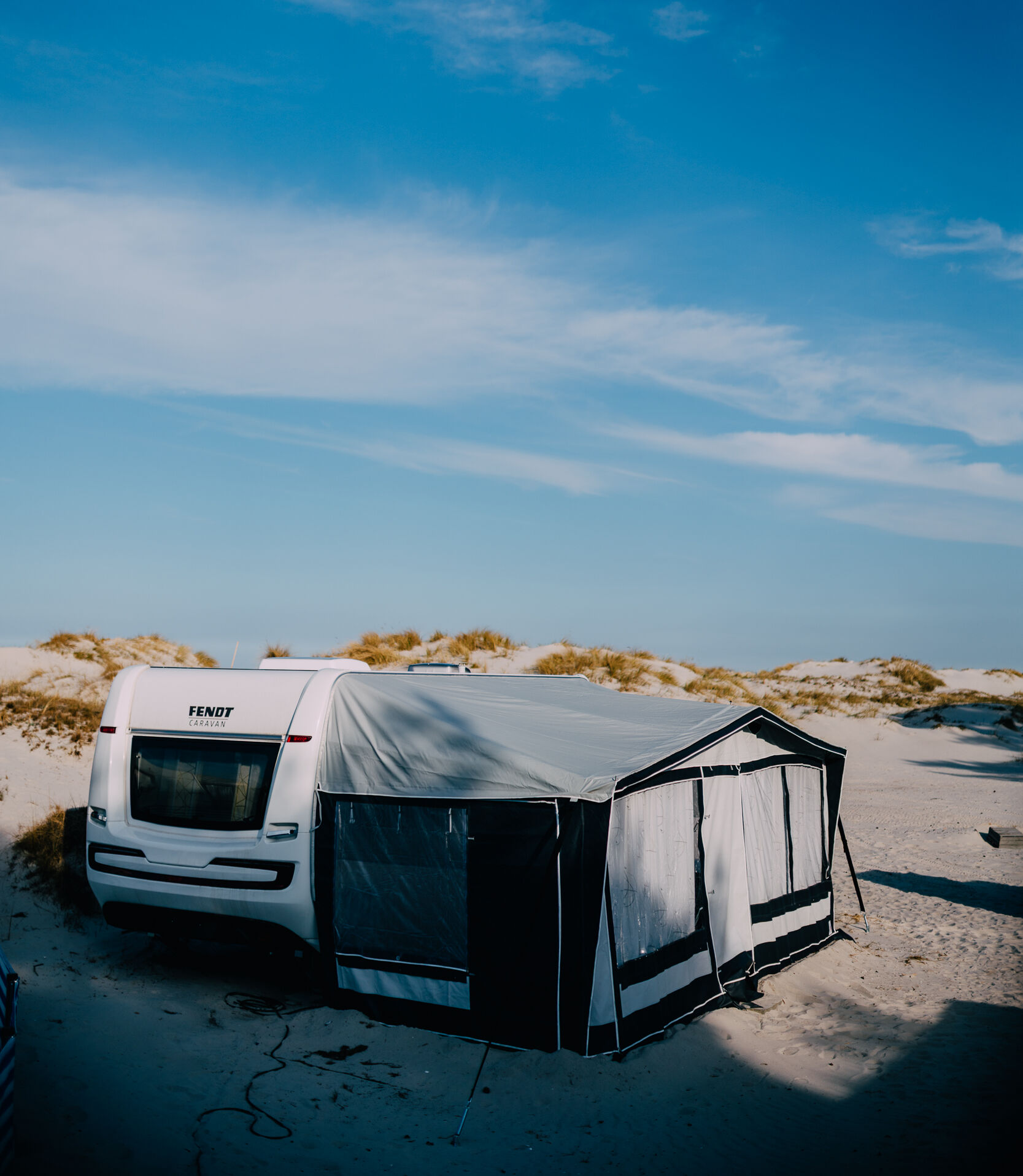 Ein weißes Wohnmobil mit angebauter Markise steht auf sandigen Dünen – ideal für Herbstcamping unter blauem Himmel.