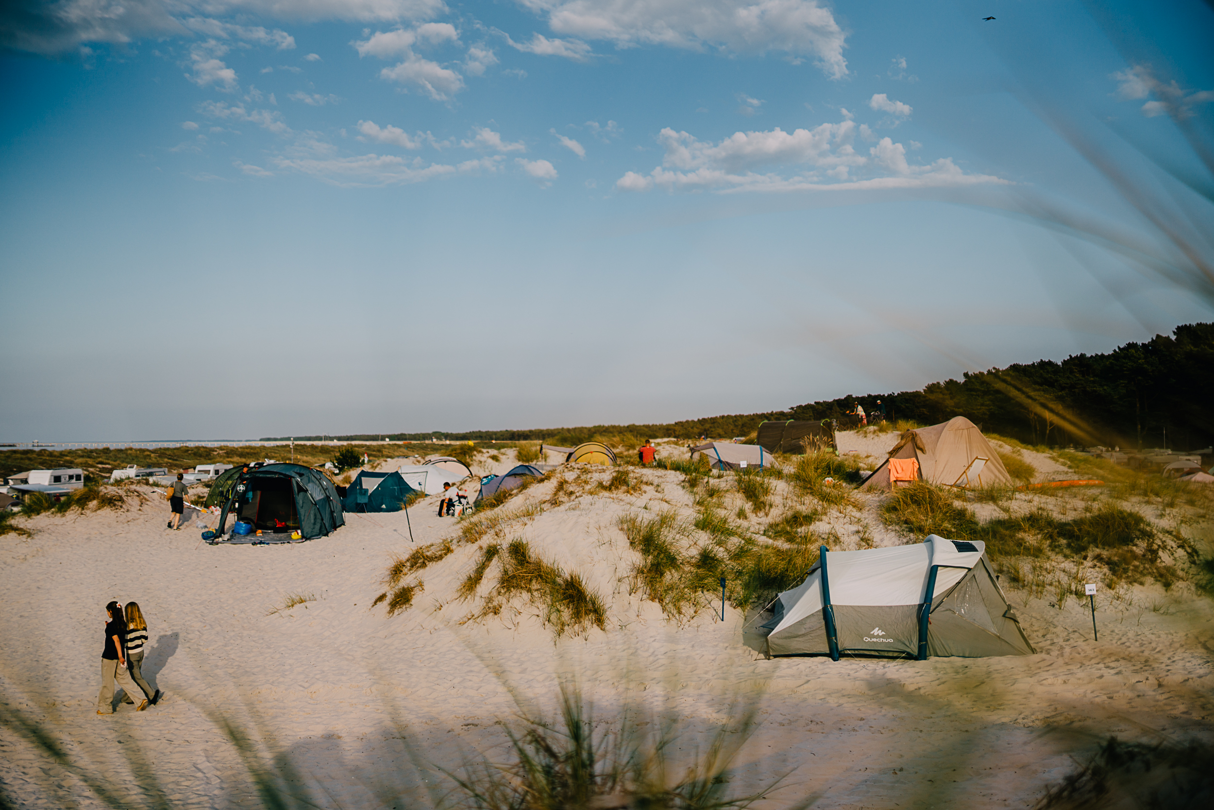 Campingplätze mit Zelten auf Sanddünen; zwei Menschen spazieren unter blauem Himmel am Strand entlang und genießen die Freiheit der Natur, nachdem sie sich für einen Campingplatz entschieden haben, den sie online gebucht haben, um ihren perfekten Urlaub zu verbringen.