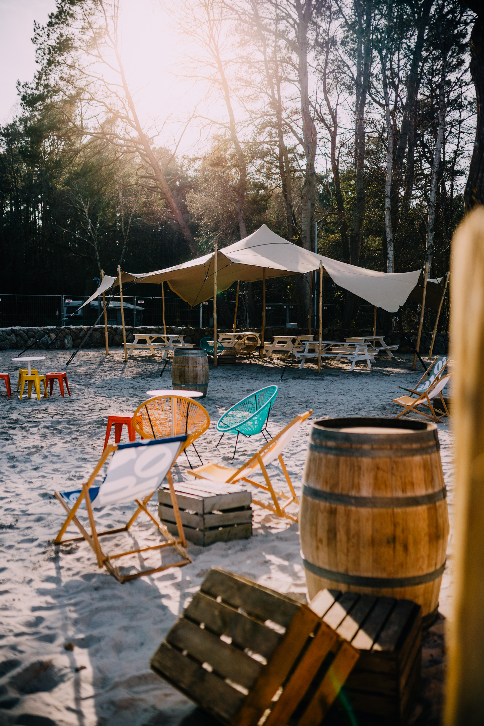 Sitzgelegenheiten im Freien am Strand mit bunten Stühlen, Holzfässern, Picknicktischen und einem Vordach, die Sonne scheint durch die Bäume.