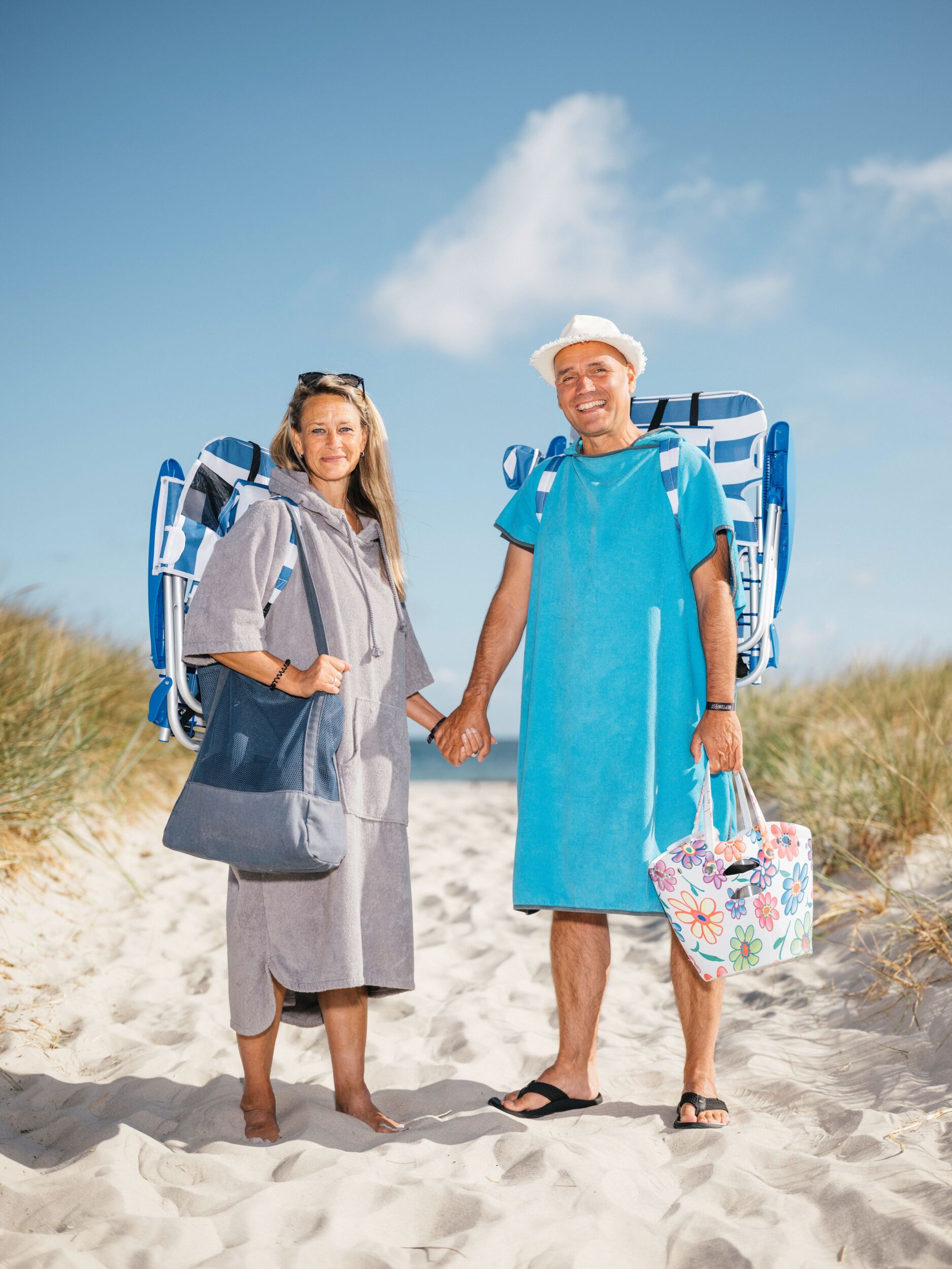 Zwei Personen in Strandkleidung, die sich an den Händen halten und Strandkörbe und Taschen tragen, stehen auf einem Sandweg mit Gras.