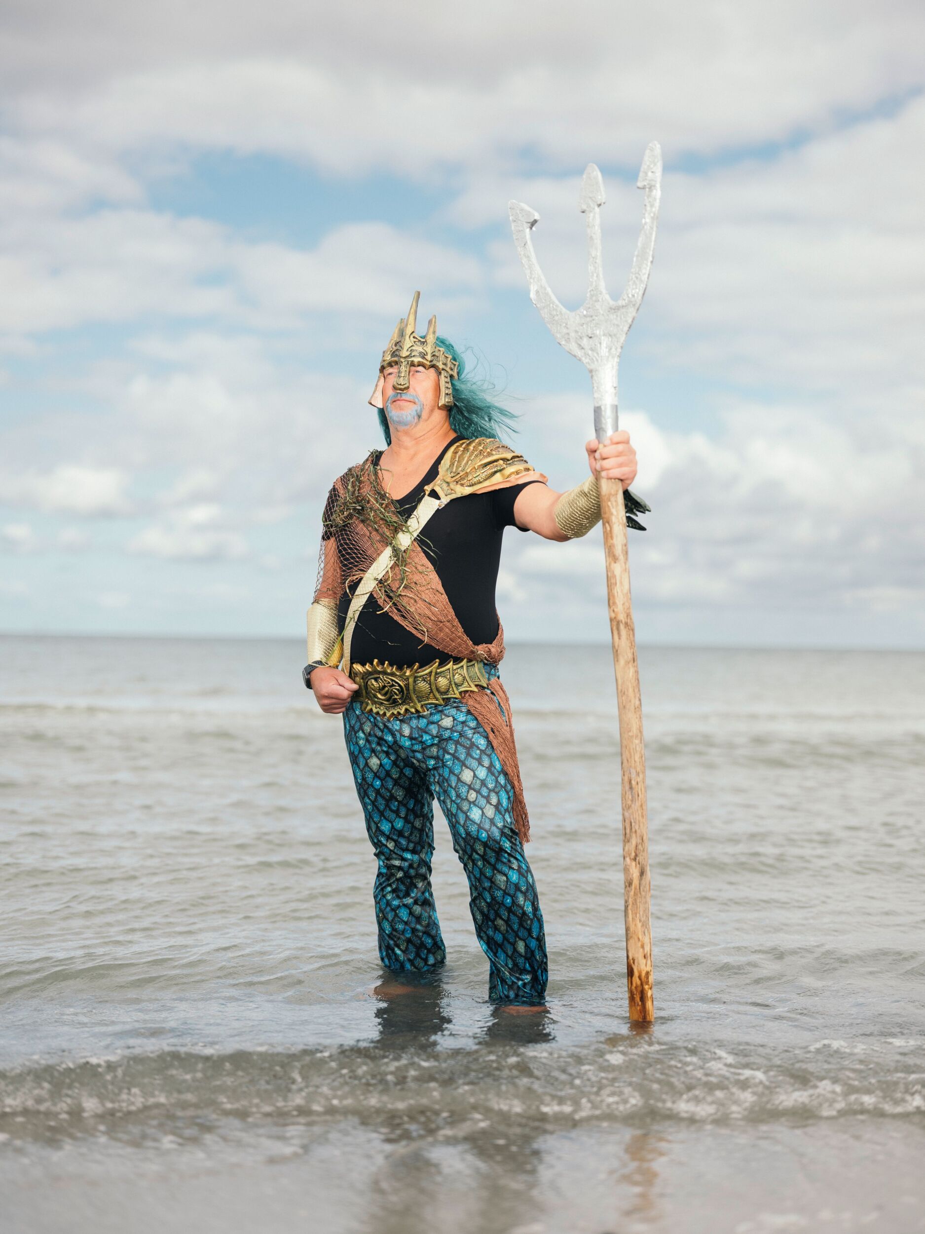 Eine als Meereskönig gekleidete Person mit einem Dreizack steht im flachen Wasser des Ozeans unter einem bewölkten Himmel.