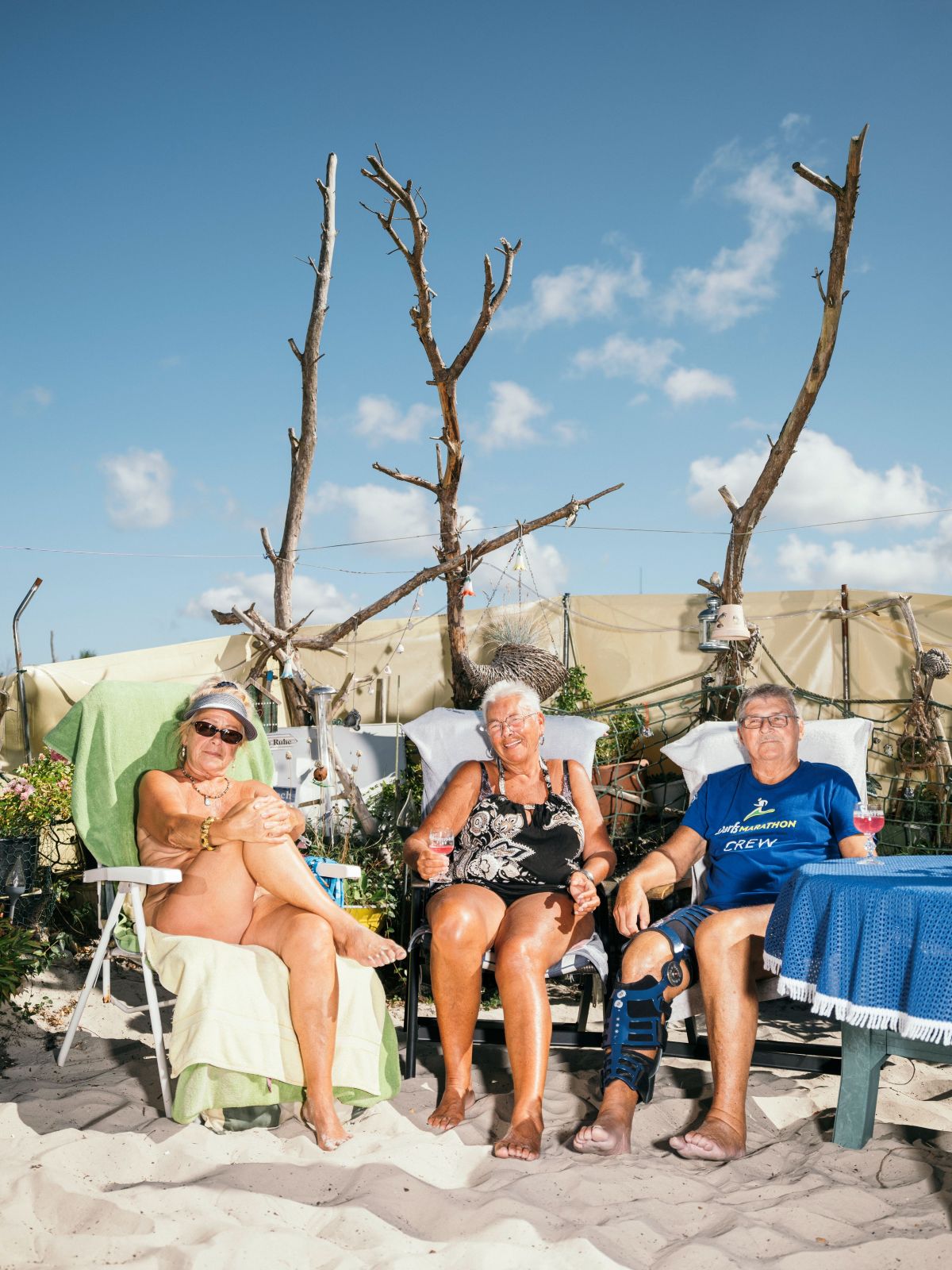 Drei ältere Erwachsene entspannen sich an einem Sandstrand mit Getränken, umgeben von behelfsmäßiger Holzdekoration und blauem Himmel.