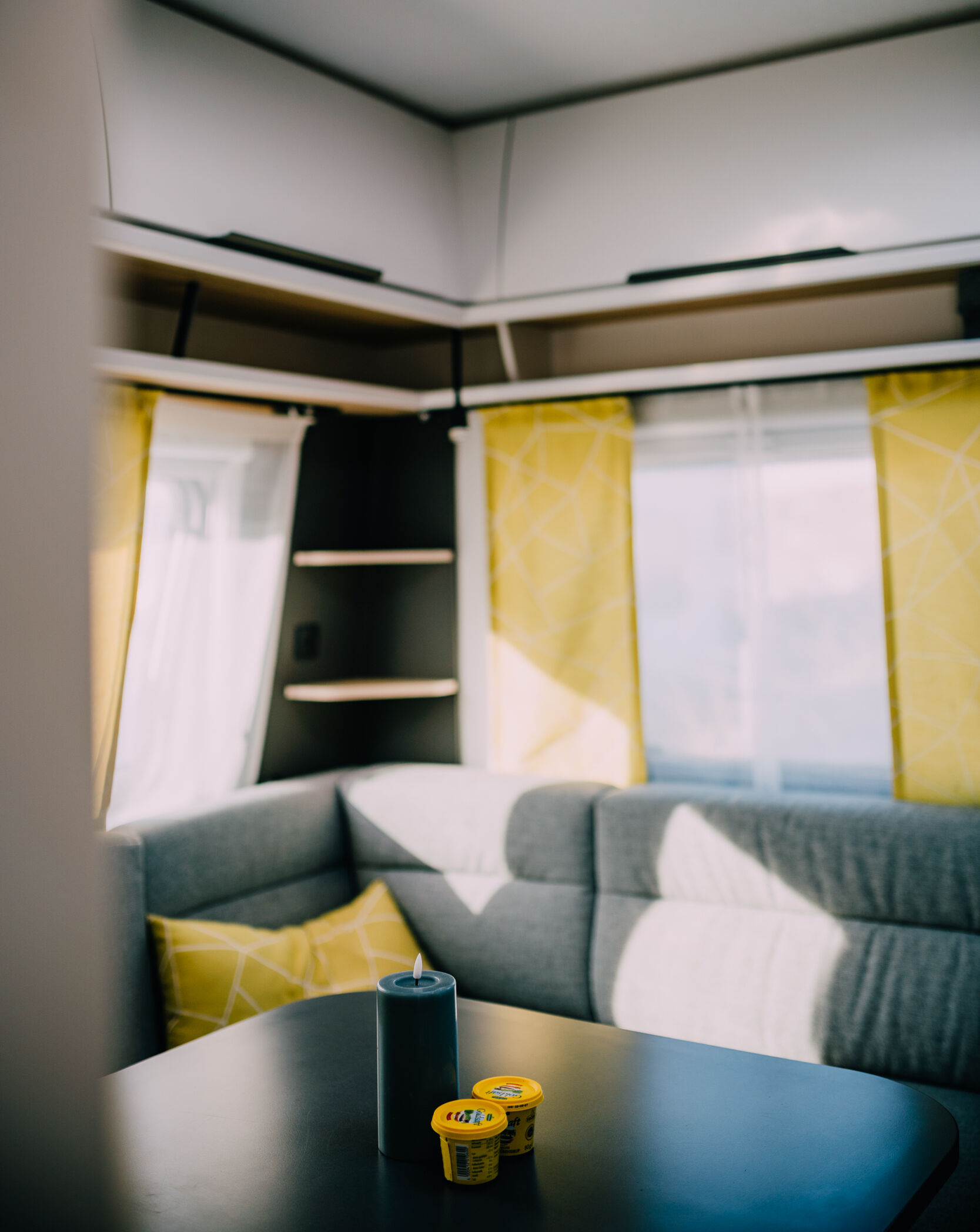 Modernes Wohnwagen-Interieur mit gelben Vorhängen, grauem Sofa und Gegenständen auf einem schwarzen Tisch bei natürlichem Licht - die perfekten Mietunterkünfte zum Entspannen nach einem Tag in den Dünen.