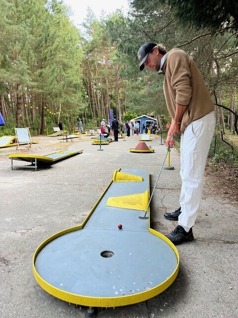 Eine Person spielt im Freien Minigolf und zielt auf ein Loch auf einem ungewöhnlich gestalteten Platz, der von Bäumen umgeben ist.